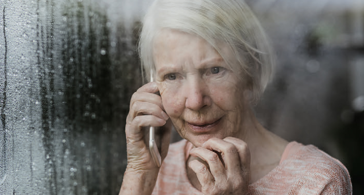 Za szybą po której spływają krople deszczu stoi starsza kobieta, która rozmawia przez telefon.