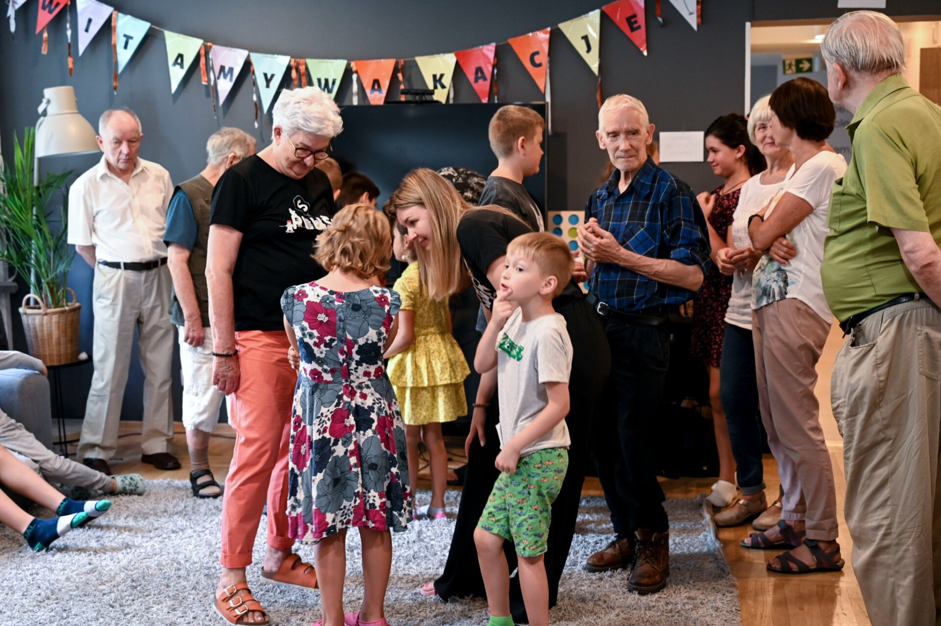 Grupa seniorów i dzieci w różnym wieku stoi w sali ozdobionej girlandą.