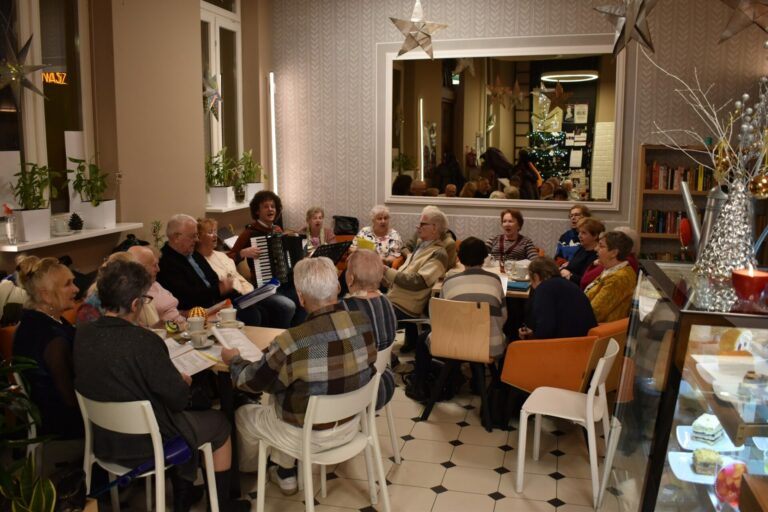 Zdjęcie przedstawia kilkanaście osób w przestrzeni kawiarnianej, śpiewających kolędy.