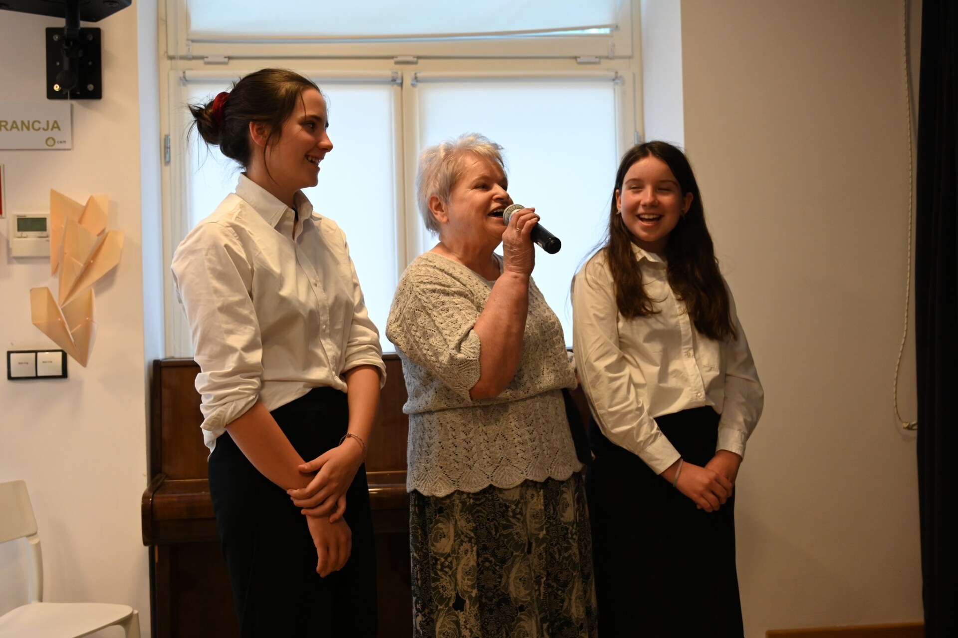Dwie młode kobiety i seniorka śpiewają piosenkę. Seniorka trzyma mikrofon.