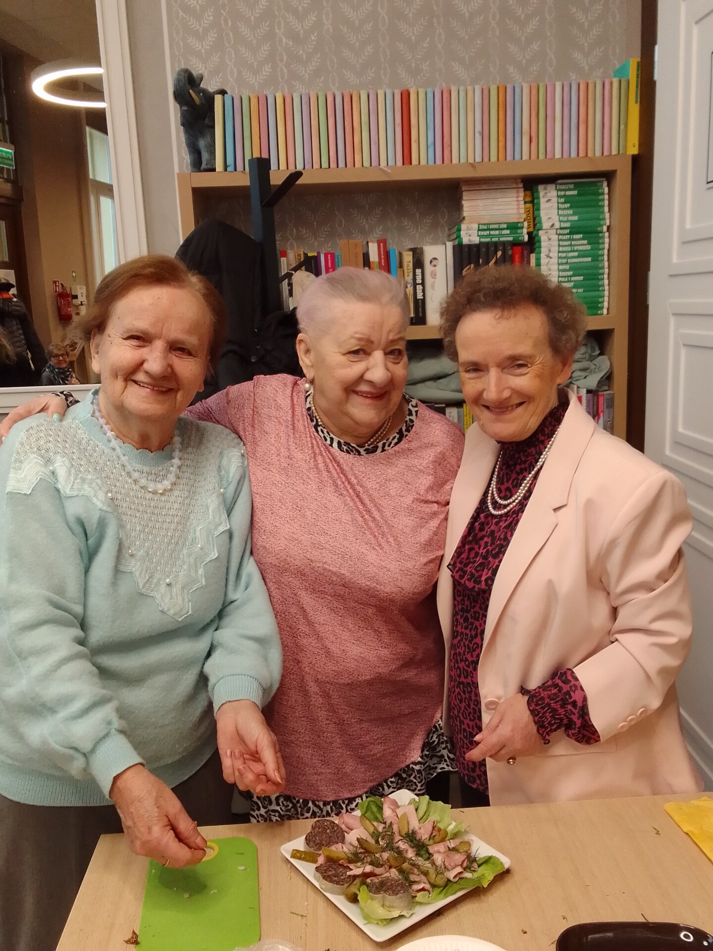 Trzy uśmiechnięte kobiety 60+ stoją przy stole na którym stoi talerz z wędlinami
