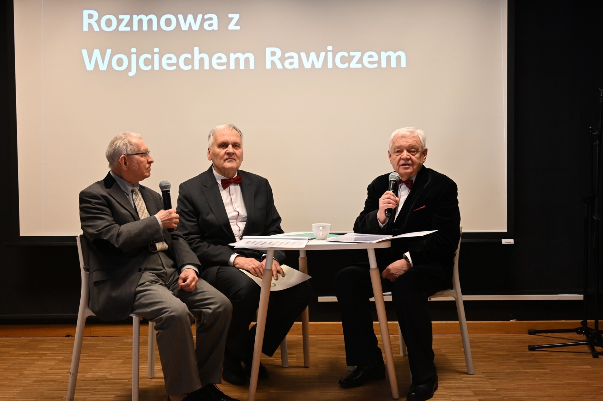 Trzej seniorzy siedzą na scenie przy stoliku, w tle napis "Rozmowa z Wojciechem Rawiczem"