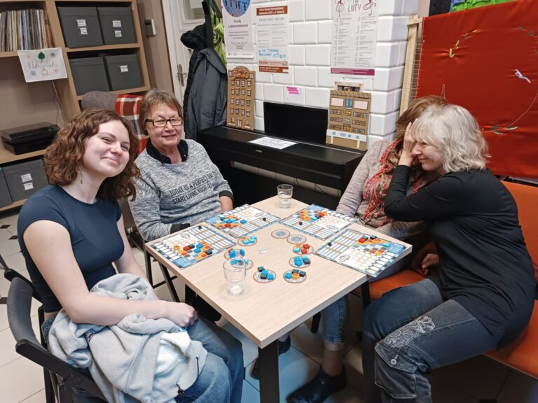 cztery kobiety siedzą przy stoliku, są uśmiechnięte i grają w grę planszową