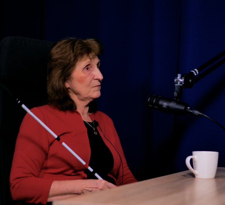 Zdjęcie przedstawia kobietę siedzącą przy stole i mówiącą do mikrofonu.